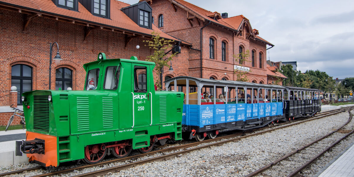 Skład wąskotortowy kolei turystycznej w Pleszewie stojący przed dworcem. Lokomotywa oraz dwa wagony. W wagonach pasażerowie jadący do Kowalewa, głównie dzieci.