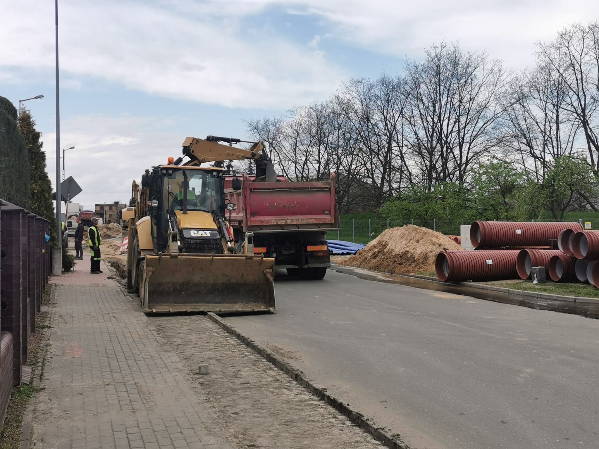 Zdjęcie przedsatwia drogę, na której znajduje się sprzęt budowlany(ciężarówka i koparka), obok leżą rury do wodociągu, stoją robotnicy